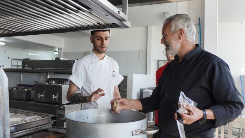 Chef y ayudante cocinan en cocina industrial junto a una olla grande.