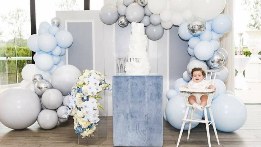 Bautizo en tonos azules: arco de globos y bebé en trona junto a tarta.