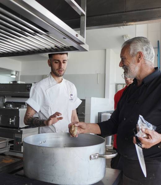 Chef y ayudante cocinan en cocina industrial junto a una olla grande.